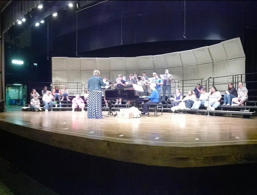 A group of singers stand and sit on risers on a stage; in front of them is a piano, an accompanist, and a choral director.