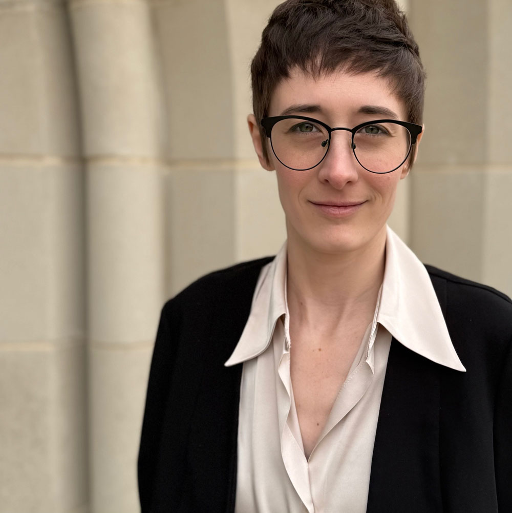 Professional headshot of a white woman with short brown hair. She is wearing glasses and smiling gently, looking directly into the camera.