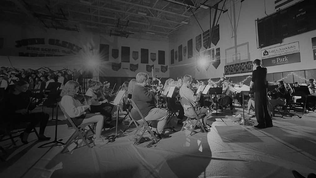 An orchestra plays while a conductor stands before the performers, arms raised. The performers are seated in a high school gym with two bright spot lights shining toward the camera. The photo is black and white.