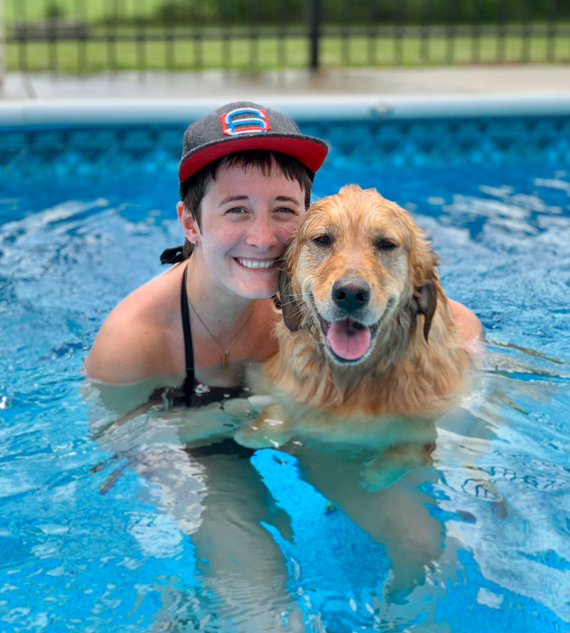 A woman with short hair holds a golden retriever; both are in a swimming pool and are smiling at the camera.