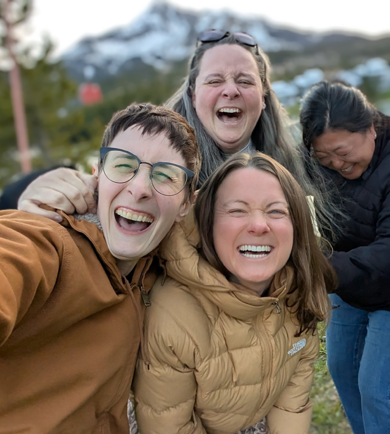 Photo of four women laughing and smiling brightly; behind them is a mountain.