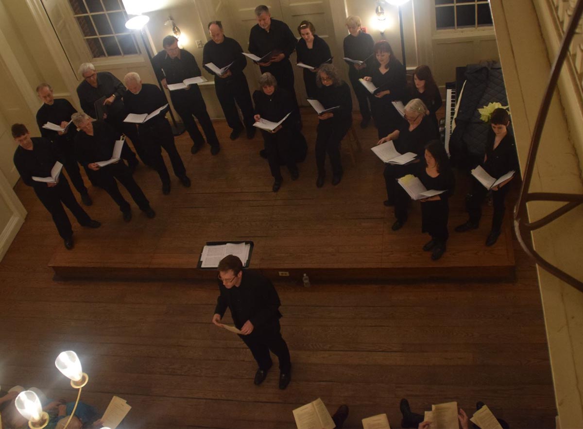 A photograph of a small choir standing in a semi-circle shot from a balcony overloooking the choir from above.